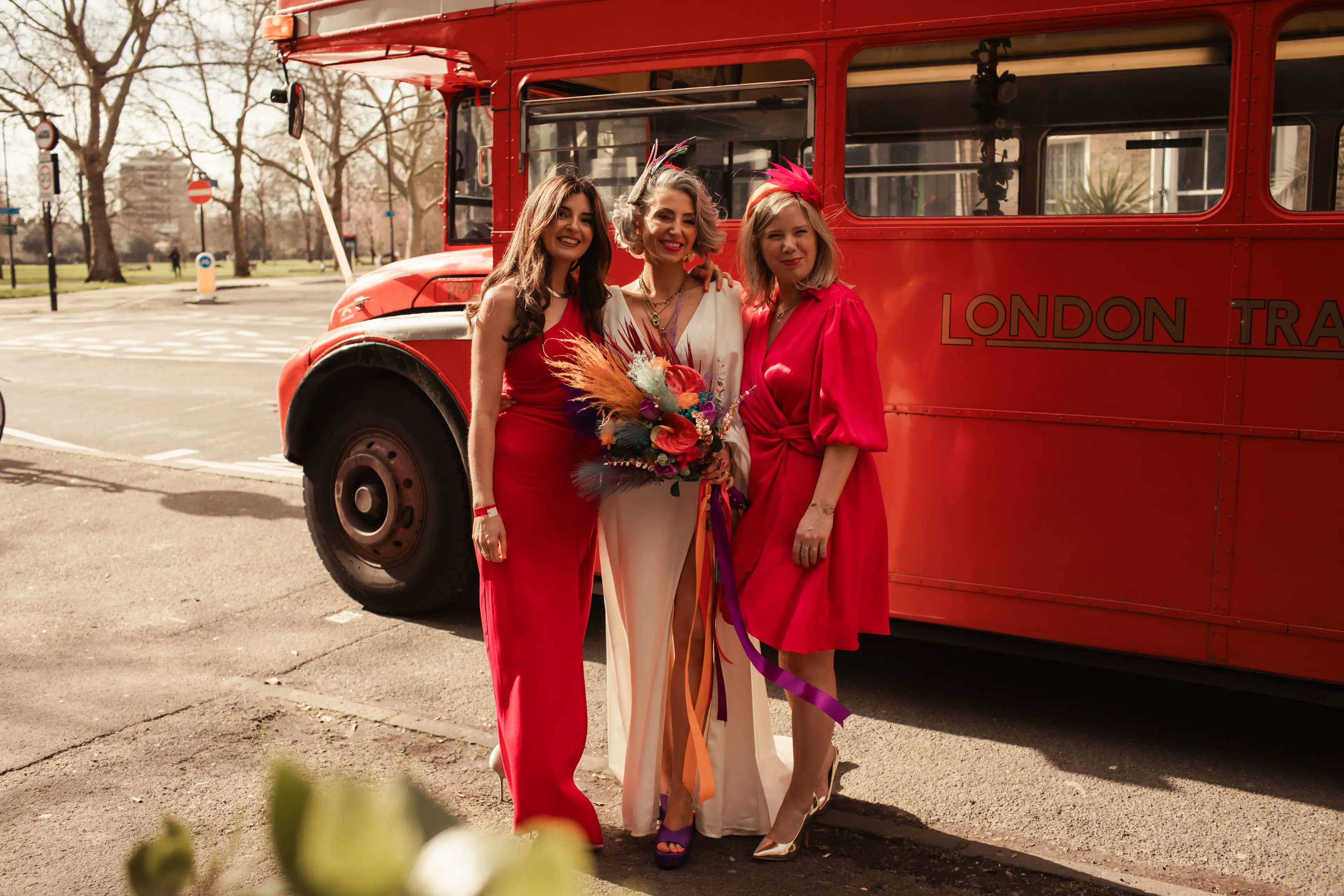 A colourfully dressed bride and some guests in front of a red London bus